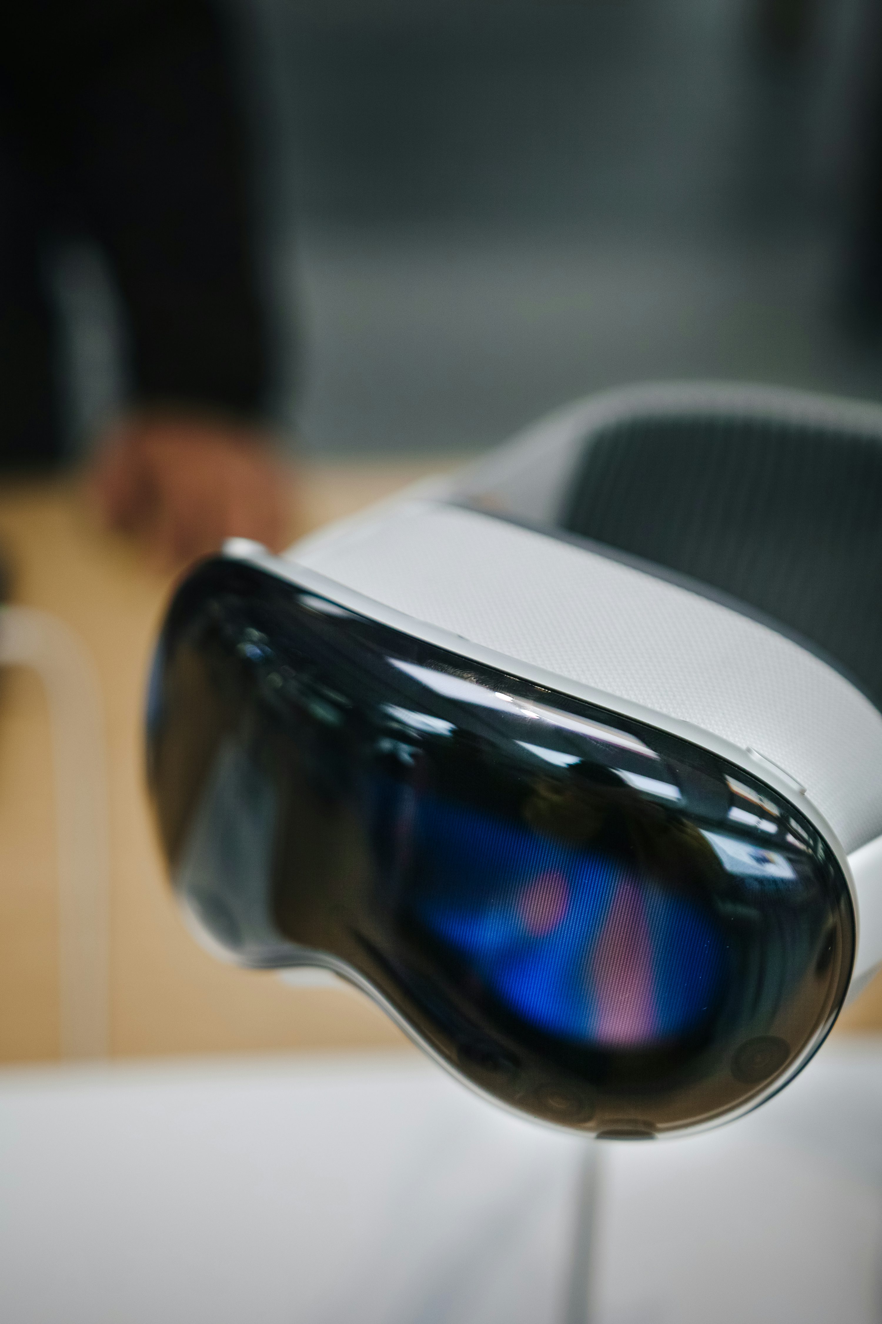 a closeup shot of a white vr headset with a sleek, modern design. the headset features a reflective black visor and a visible screen with blue and pink colors inside the visor. it is resting on a lightcolored surface, possibly a display stand or table. the background is slightly blurred, suggesting a retail or showroom setting.
