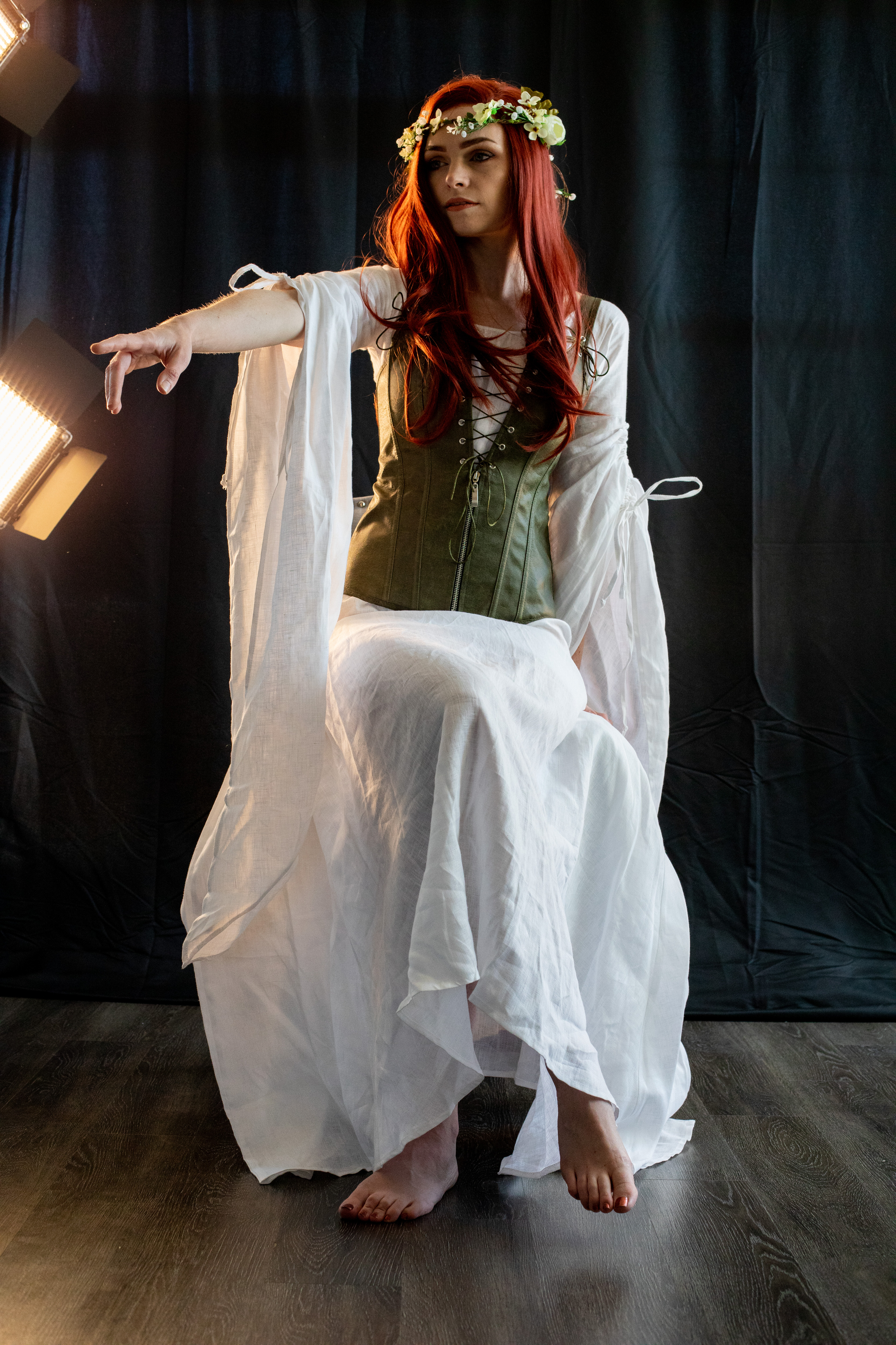 a young woman with long red hair and a flower crown poses in a medievalinspired costume. she wears a white flowing dress over a green bodice, giving the impression of a fantasy character like a princess or elf. the setting appears to be a studio, with professional lighting illuminating the subject against a dark backdrop.  she sits on a stool mostly obscured by her dress, extending one hand outward in a dramatic gesture.
