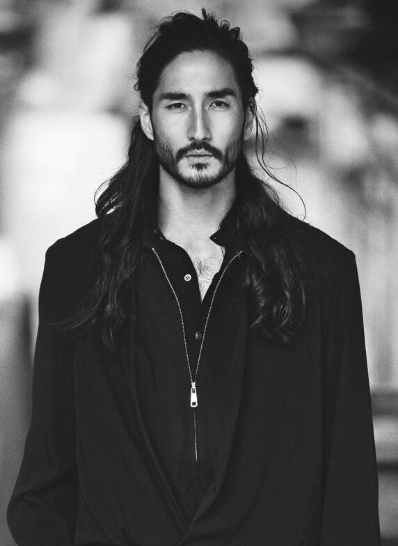 a black and white portrait of a man with long hair and a beard, wearing a dark colored shirt or jacket with a zipper.