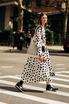 a woman wearing a black and white polka dot dress, a beige tote bag, black sneakers, and walking in a crosswalk with a city background.  she is walking down the street in a trendy outfit.