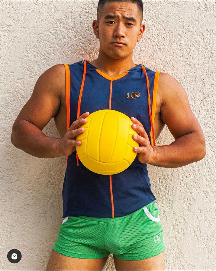 a young, muscular asian man is posing against a white wall. he is wearing a dark blue sleeveless athletic shirt with an orange trim and a small logo, green athletic shorts, and is holding a yellow volleyball. he has a serious expression on his face. the image is welllit and clearly shows the man's physique.  the overall tone is sporty and stylish.