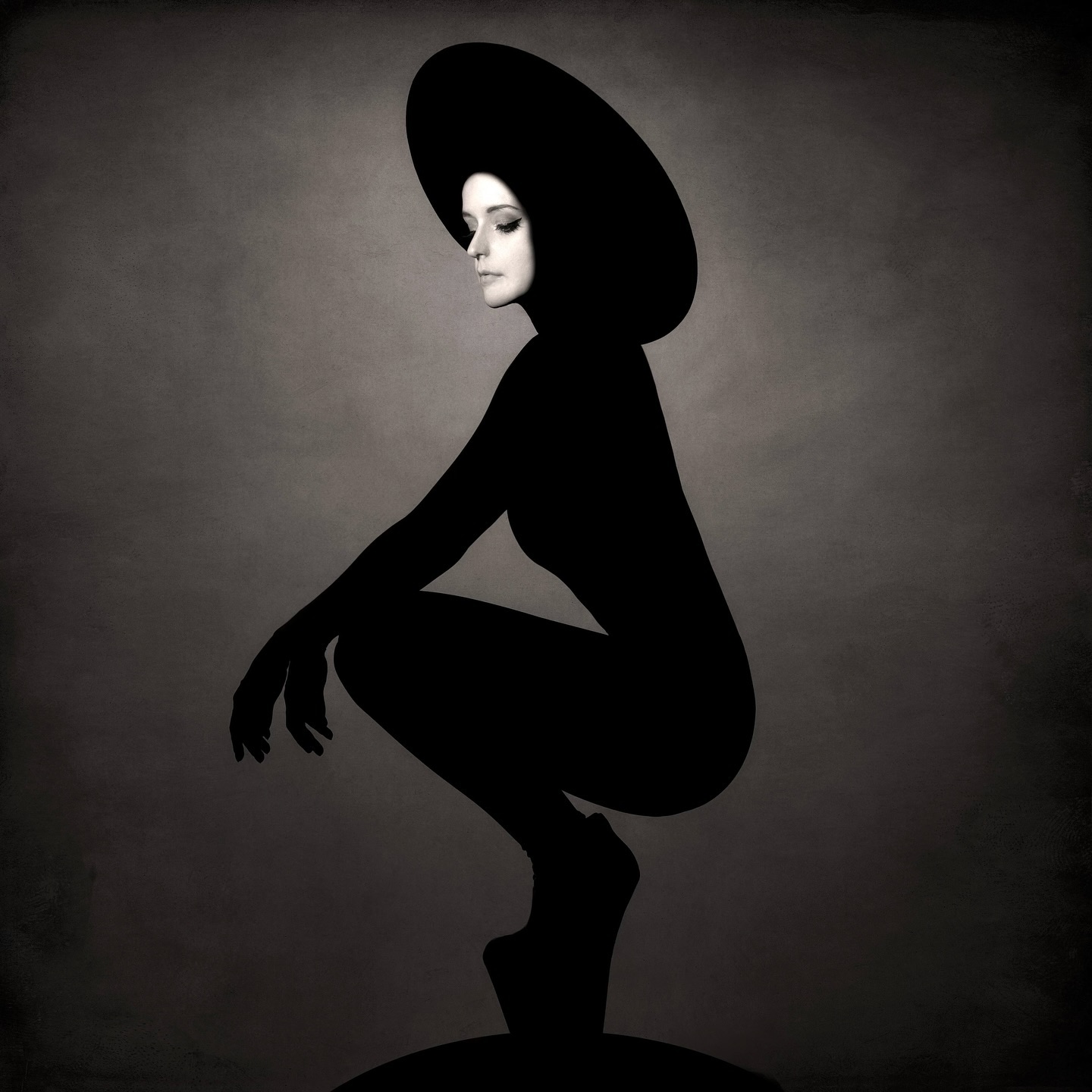 a black and white studio portrait of a woman wearing a widebrimmed hat, posing in a dramatic silhouette. she is sitting in a squat position with her legs crossed and her head slightly tilted down. the lighting is soft and moody, casting shadows that add to the mysterious and elegant feel of the image.