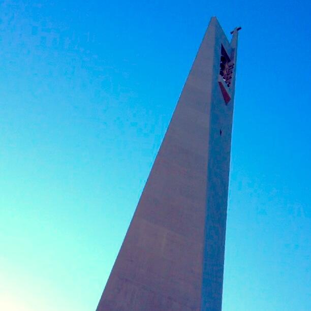 the image shows a tall, narrow, triangularshaped structure, likely a church steeple, reaching towards a bright blue sky. the perspective is from the base looking straight up.  the structure appears to be made of concrete and has a minimalist design.