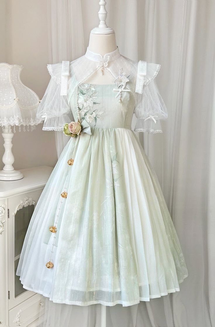 a flowy white and green vintage or lolita style dress is displayed on a headless mannequin. the dress has a floral print, lace overlay and tulle sleeves. there is a white cabinet to the left and a white wall in the background.
