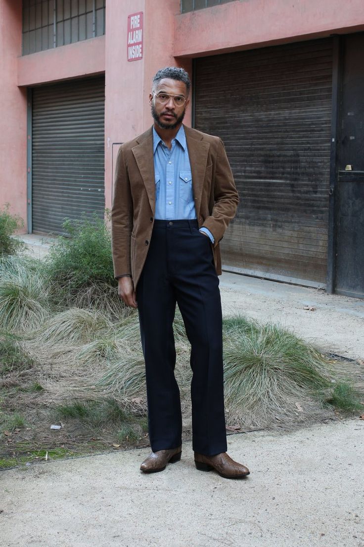 a fullbody shot of a man standing outdoors, in front of a building. he is wearing a brown corduroy jacket, a light blue buttondown shirt, dark trousers, and brown boots. his style is sharp and put together. the setting is urban, with visible building walls and grassy patches.  the man appears to be stylishly dressed.
