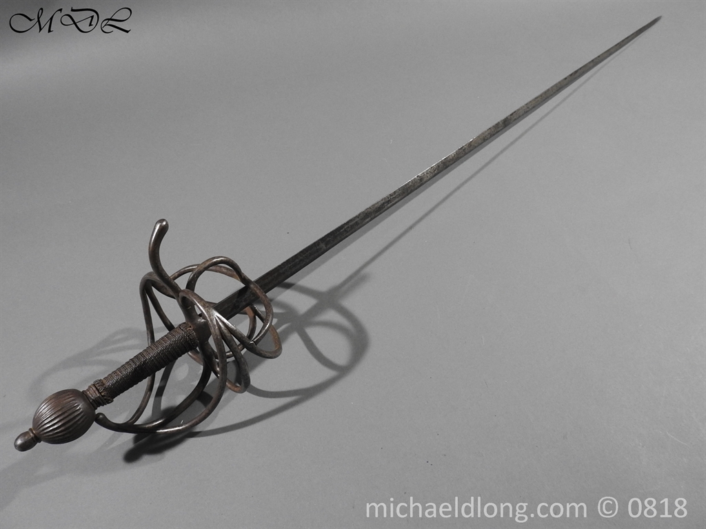 an antique sword lays on a gray background. the sword is pointed towards the upper right corner of the image.  the hilt is a complex basket weave of metal designed to protect the hand. the pommel at the end of the grip is round.