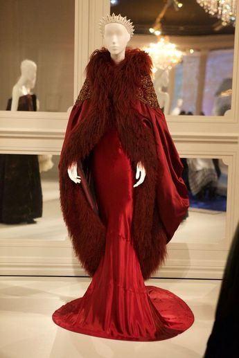 a mannequin is wearing a stunning red gown and fur cape. the gown is fitted and has a long train. the cape is made of dark brown fur and has a collar. the mannequin is wearing a crown and has a neutral expression on her face. she is standing in a museum, and the floor is polished. the gown has a lot of details, and it is clear that this is a highend fashion piece.