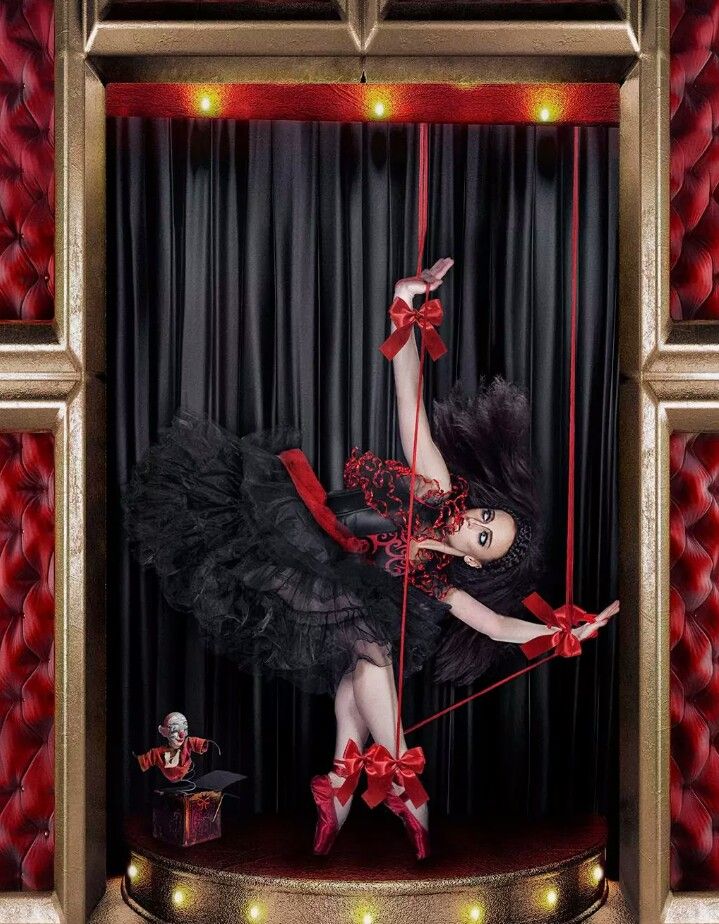 a woman in a black dress and red shoes is being manipulated like a puppet by red strings. she is on a stage in a theatre with red velvet curtains in the background. the image is taken from a unique perspective, giving the viewer a view from above.