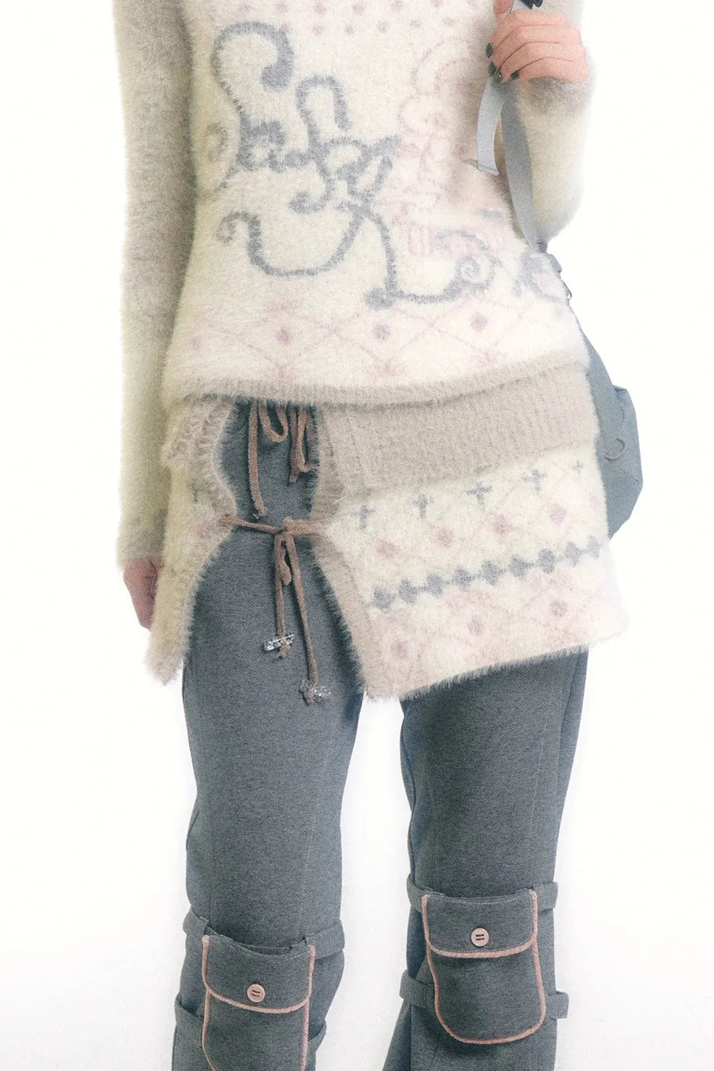 loseup view of a woman's outfit.  she's wearing a fluffy, light beige sweater with a pale pink and grey pattern. over the sweater, she has on a layered, similarly textured skirt, also in beige and pink hues, that has a unique asymmetrical, wraparound design and is tied with cords. underneath the skirt are dark grey pants with added pockets on the legs, secured with straps.  the overall style is layered, soft, and somewhat whimsical. the image is a studio shot on a white background.