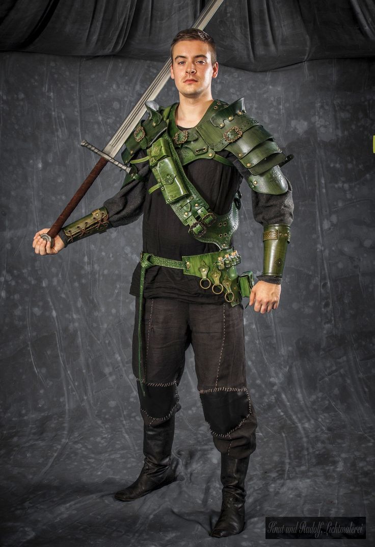 a fullbody studio shot of a man in a medieval fantasy costume. he is wearing green leather armor, including pauldrons, a chest piece, gauntlets, and a belt with a quiver. he is also wearing dark pants and boots. he is holding a large sword, which rests across his back. the background is a dark grey fabric backdrop. the image is welllit and sharply focused.