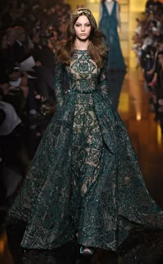 a woman in a beautiful green gown with intricate embroidery and lace details walks down the runway during a fashion show. the dress is fulllength and features a fitted bodice and a flowing skirt. the model's hair is styled in a sleek, straight look, and she is wearing minimal jewelry. the runway is a dark surface, and there is a blurred background of people watching the show.