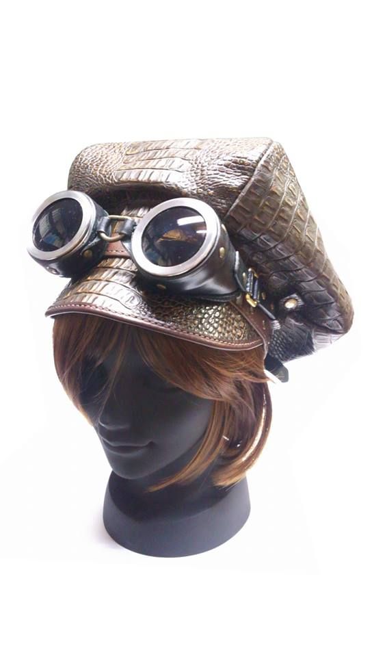 a closeup shot of a brown crocodile leather hat with goggles attached to it. the hat is sitting on a mannequin head with brown hair. the hat is a stylish and unique accessory that would be perfect for a steampunk or vintage look.