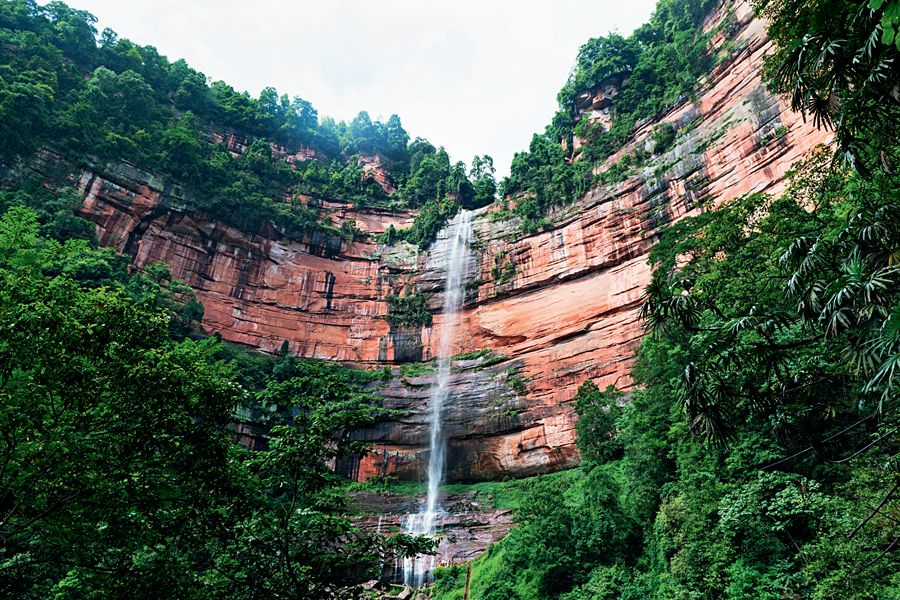 the image showcases a tall waterfall cascading down a steep, layered red rock cliff face. the cliff is densely covered with green trees and vegetation at the top and base, creating a lush, natural environment. the waterfall itself is a prominent white stream against the colorful rock, surrounded by the vibrant greenery. the view captures a picturesque and serene scene of nature's beauty.