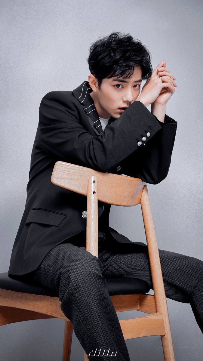 a young man with styled black hair is wearing a black suit jacket with white pinstripes and matching pants. he is seated on a wooden chair with his hands clasped in front of his face, his expression serious and engaging with the camera. the background is a solid light gray, creating a minimalist and studiolike setting. the lighting is soft, casting subtle shadows and highlighting the textures of his clothing.
