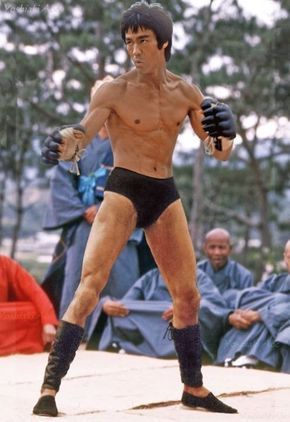 bruce lee is shown striking a fighting pose, with his arms extended and fists clenched, ready to strike. he is wearing black shorts and black boots, and is shirtless to showcase his muscular physique. the image is black and white, which adds to the vintage feel of the photograph. in the background, there is a group of people seated and observing lee, perhaps an audience watching a demonstration or a training session.  the overall image highlights lee's strength, focus and the dynamic energy of martial arts.