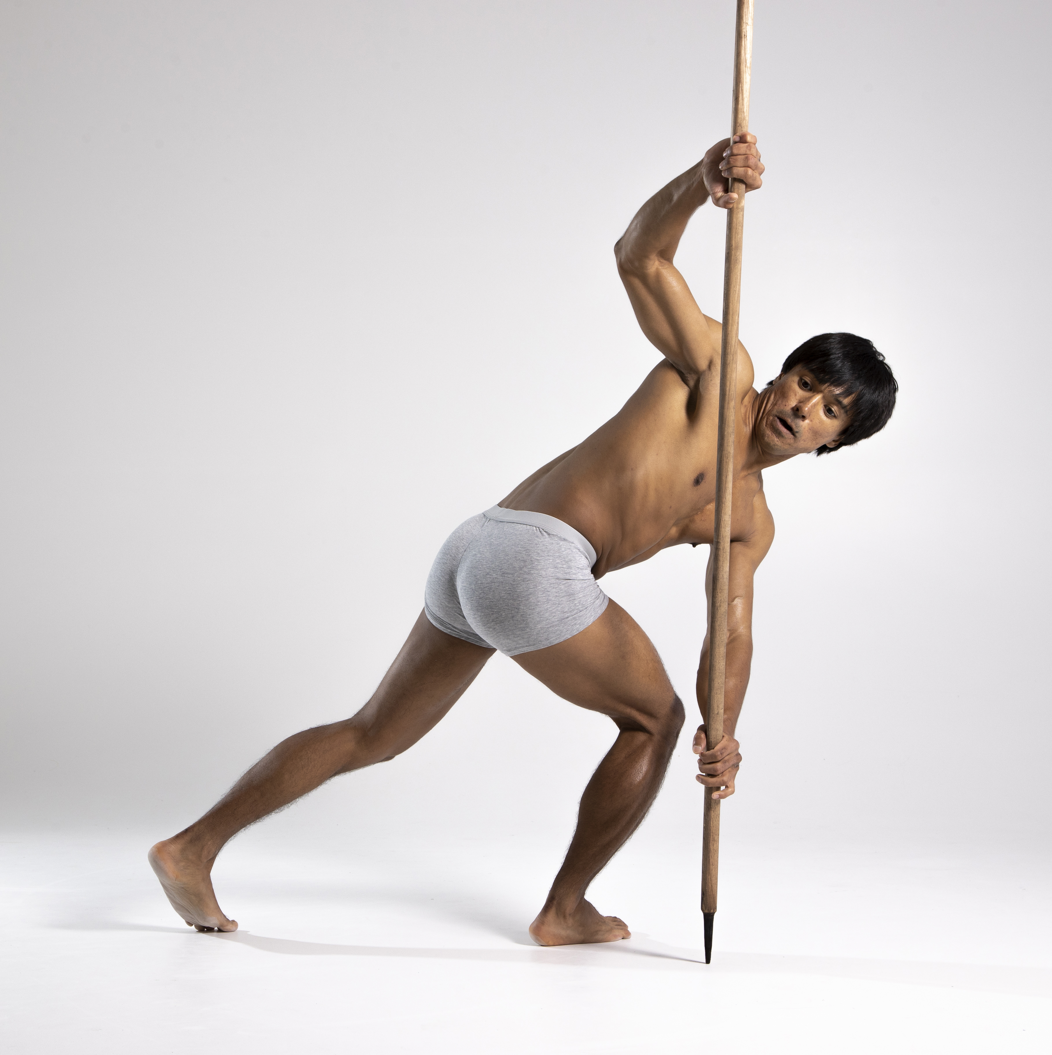 a shirtless man in gray underwear is holding a spear and leaning on it for support. he is barefoot and in an athletic pose against a plain white background.