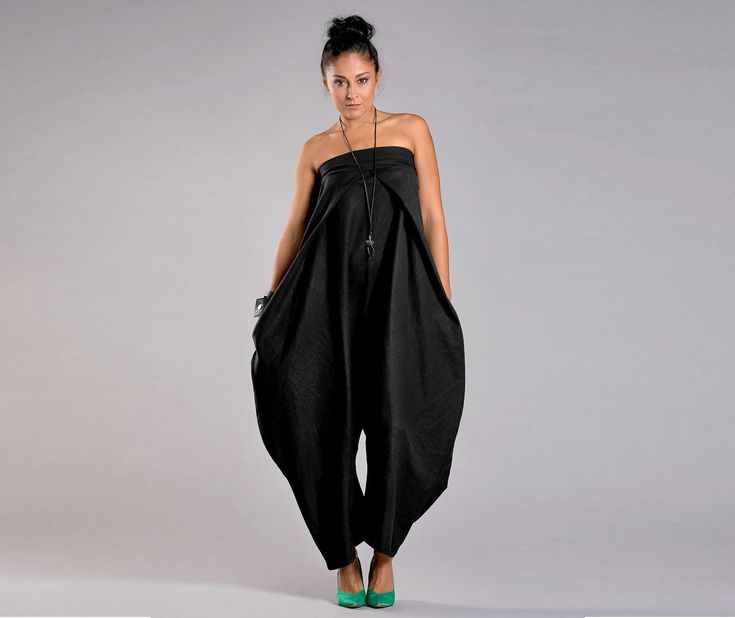 a woman wearing a black strapless jumpsuit is standing in a studio. she has her hair pulled back and is wearing green heels. the background is a light gray.
