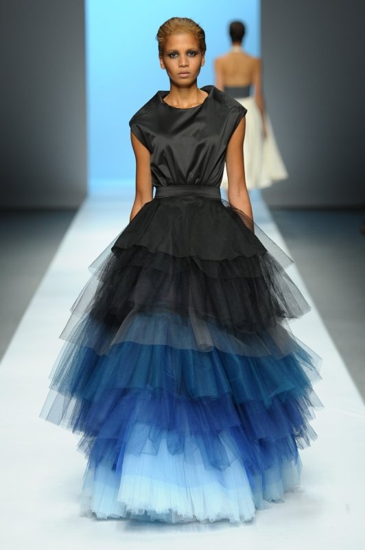 a woman is walking on a runway in a black and blue ombre dress. the dress has a black top and a tiered tulle skirt that fades from black to blue. another person is walking in the background.