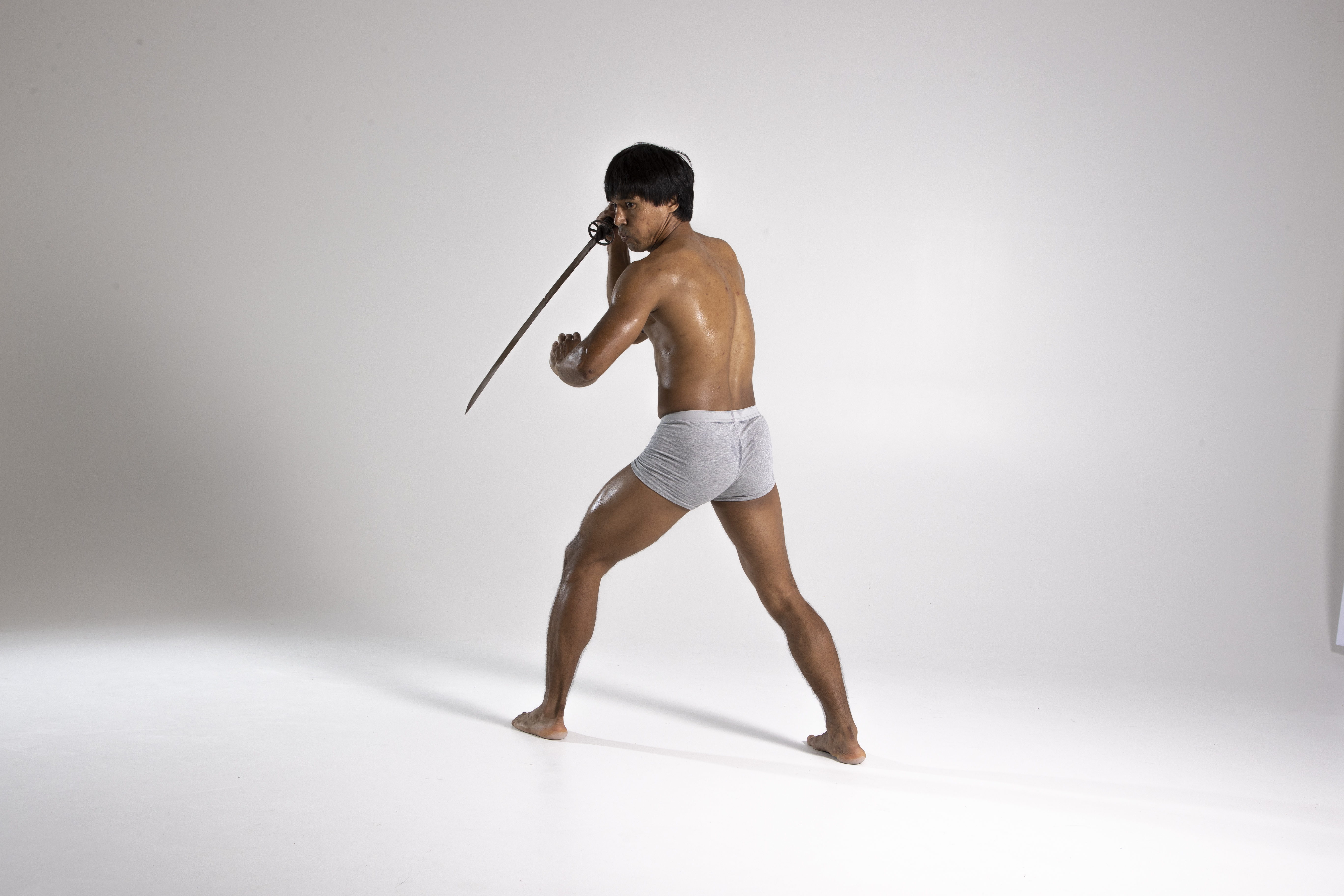 a shirtless man in grey underwear is holding a sword in a defensive pose. he is barefoot and in a fighting stance, with a slightly sweaty and muscular build. he is in a studio setting with a plain white background.