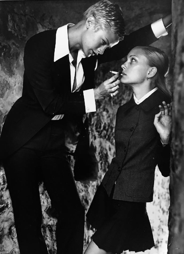 two women wearing black suits. the woman on the left is smoking a cigarette, leaning in toward the woman on the right as she takes a drag. the women are serious, staring intently at the camera.