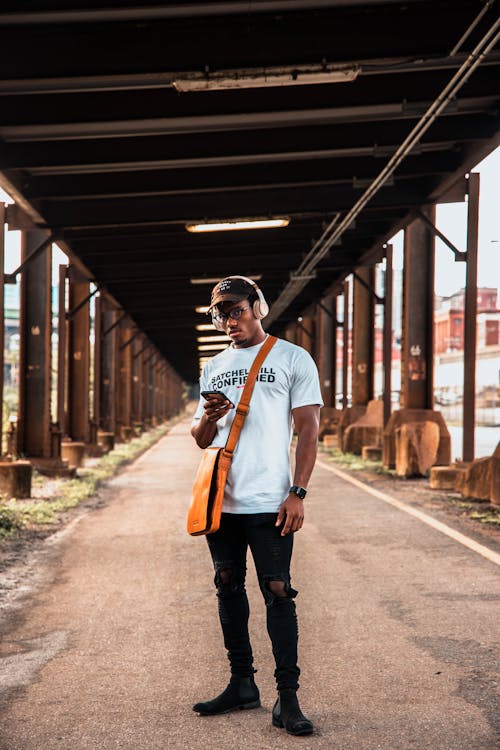 a young black man is standing in an urban underpass, looking down at his smartphone. he's wearing a white tshirt with text, black ripped jeans, chelsea boots, and a brown leather messenger bag across his body. he also has wireless overear headphones and a baseball cap on. his expression is serious and focused. the background is a long, somewhat rustic underpass with stone walls and supports. the overall mood is urban and stylish.