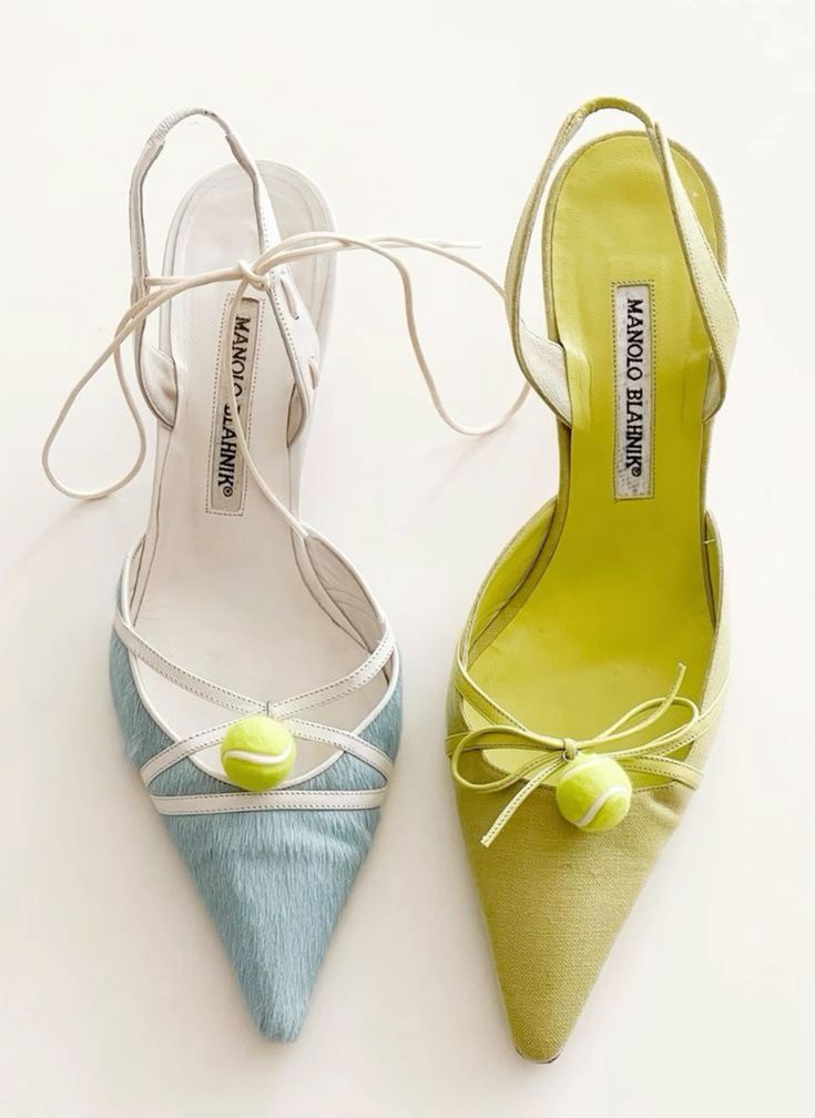 a pair of manolo blahnik heels, one blue and one green, both decorated with a small tennis ball on the toe. the shoes are both high heels and have a pointed toe. the blue shoe has a white strap across the top and a white strap around the ankle. the green shoe has a green ribbon tied around the ankle and a white ribbon across the top.