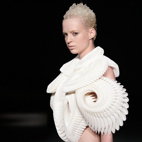 a female model wearing a white, futuristic, and geometric dress with an abstract 3dprinted design on a runway.  the dress has a sculpted and architectural look, with intricate layers of fabric and sharp angles.