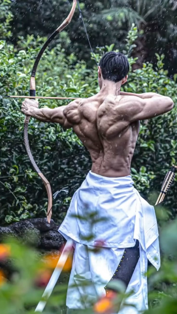 a muscular man with his back to the camera is drawing back a bow and arrow in a natural outdoor setting. he is wearing a traditional japanese hakama and has a katana sword at his side, suggesting the practice of kyudo, or japanese archery. it is raining.