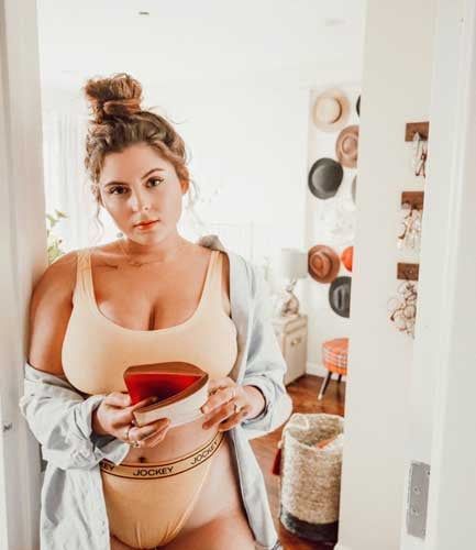 a young, curvy woman with her hair in a bun stands in a doorway, holding a red and white object (partially obscured) in her hands. she is wearing a pale yellow bra and underwear from the brand jockey, and has a light blue shirt draped over her arms. the room behind her is decorated with a boho aesthetic, featuring woven baskets, a hat wall, and a wooden stool. the image is welllit with natural light, creating a bright and airy atmosphere.