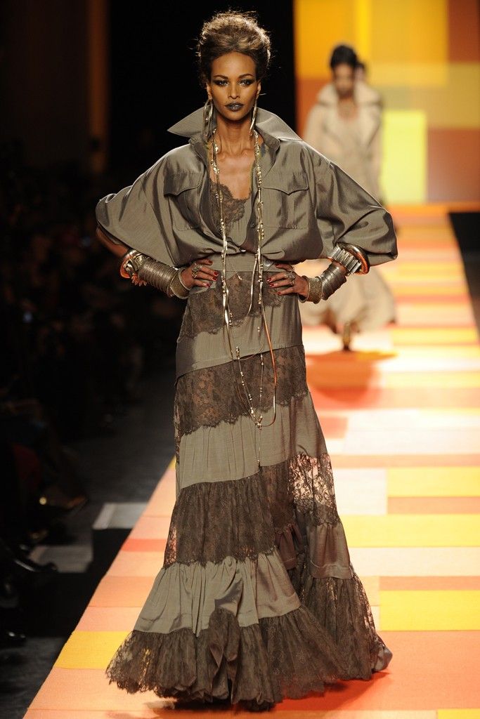 a fashion model walks down a runway in a long, flowing dress. the dress is a mix of brown silk and lace. the model has dark skin, brown hair, and is wearing silver and gold jewelry. the background of the runway is orange and yellow stripes.