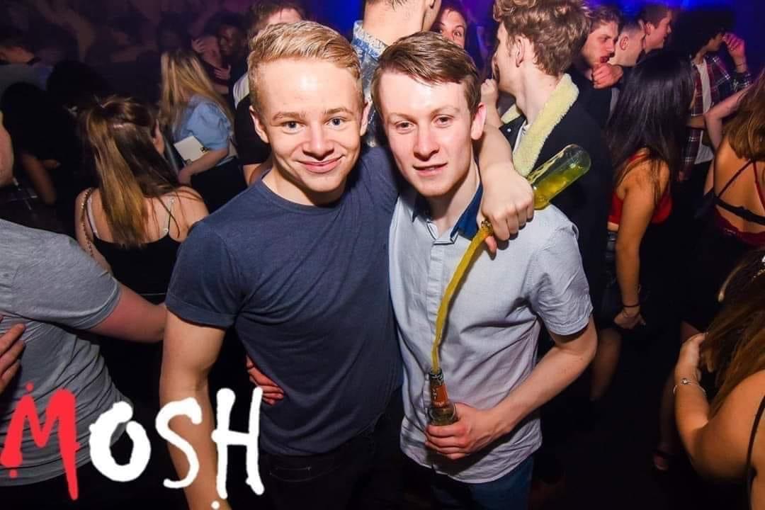 the image captures a lively scene inside a nightclub. two young men, likely friends, are the central focus. they are smiling brightly, with one arm around each other's shoulders. one is holding a beer bottle with a long straw, the contents spilling as he gestures. they are surrounded by a crowd of people enjoying themselves on a dance floor, illuminated by colorful stage lighting. the overall ambiance is one of energetic celebration and youthful revelry. the word "mosh" is superimposed on the bottom left corner, hinting at the possibly intense atmosphere.