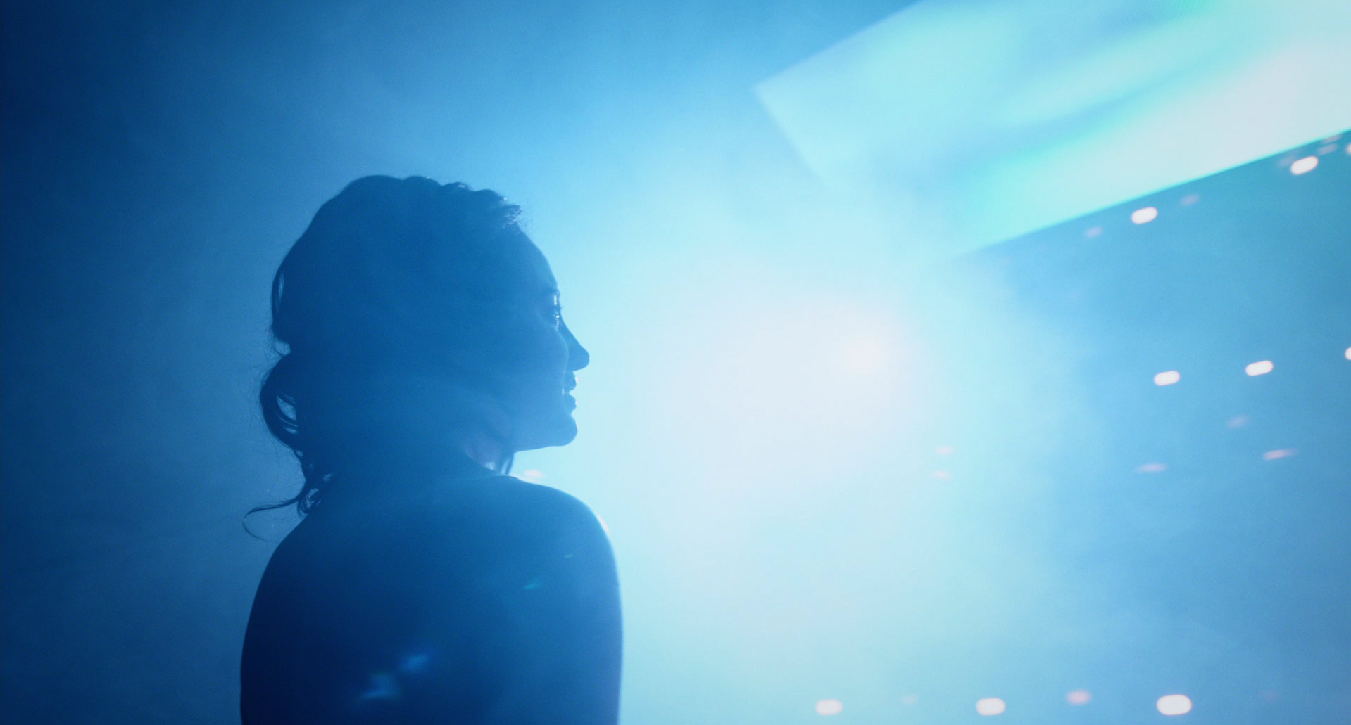 a woman is shown in profile view, silhouetted against a bright blue stage light.  the background is dark and filled with atmospheric smoke, creating a dramatic and energetic mood. circular lights are visible in the background, suggesting a concert or performance setting.