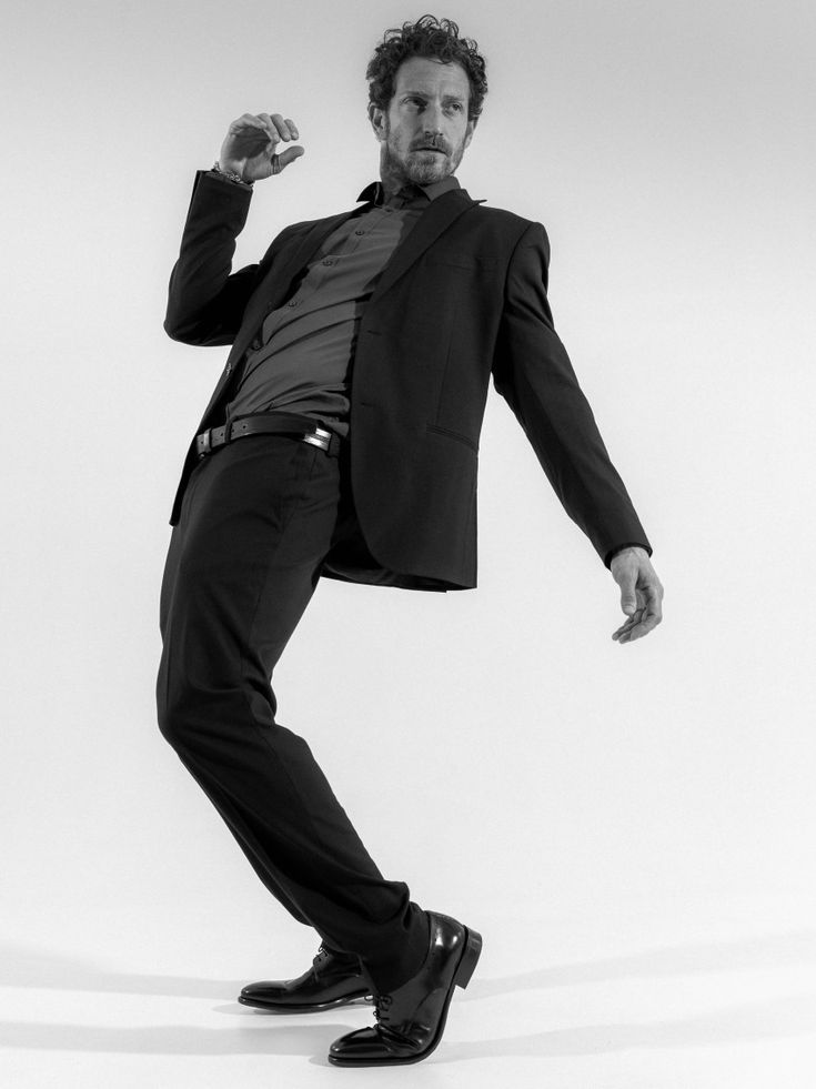 a black and white fullbody studio shot of a stylish man in a black suit and black shoes. he is posing dynamically, with one leg bent and his body angled, suggesting movement and attitude. his expression is confident and cool. the background is plain white, focusing all attention on the model and his attire. the image is sharp and clean, presenting a sophisticated and contemporary feel.