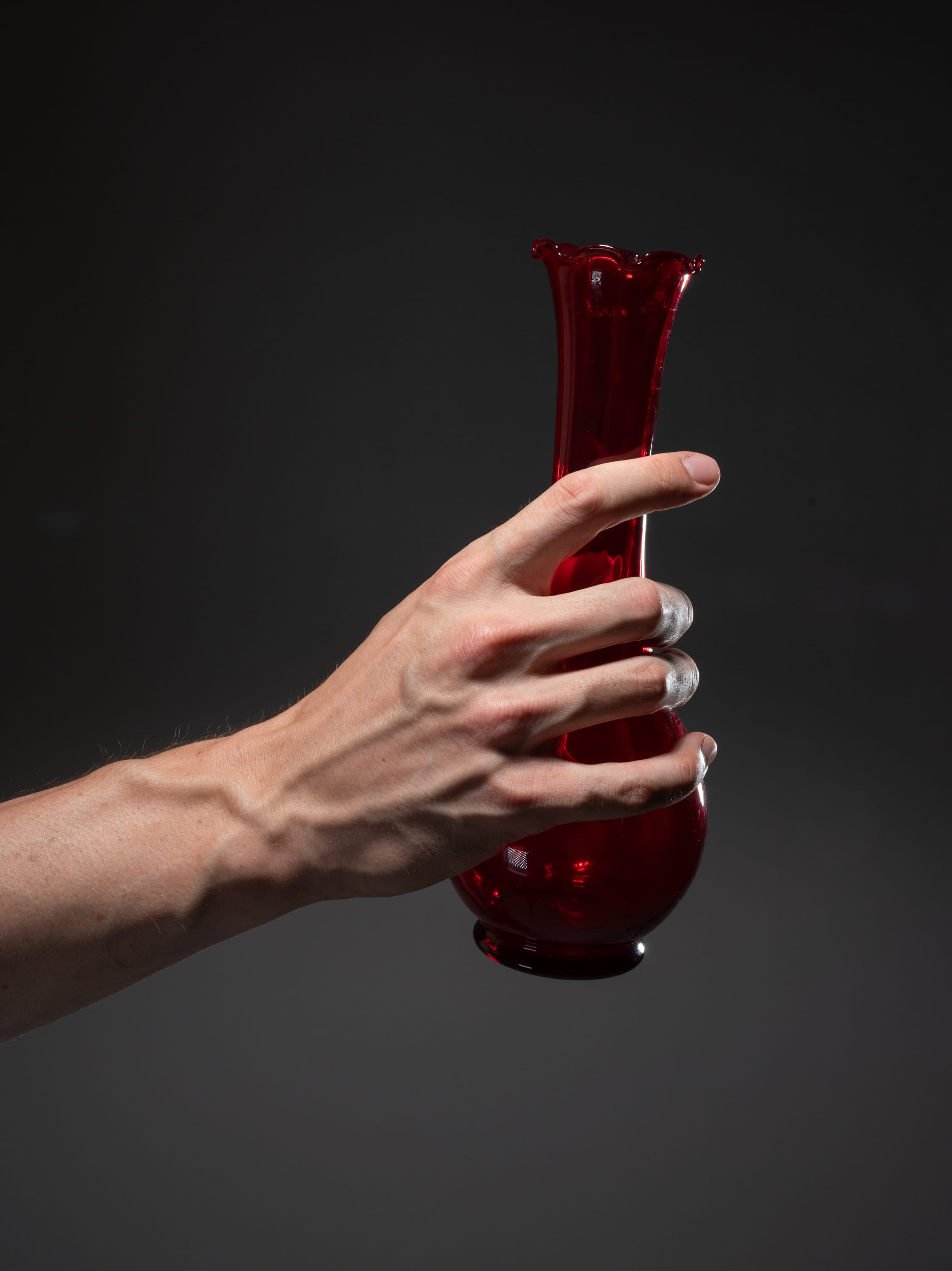 a caucasian male hand is holding a transparent red glass vase against a dark gray background. the vase has a narrow neck and a bulbous base. the hand is illuminated by studio lighting, creating highlights on the skin and the vase.