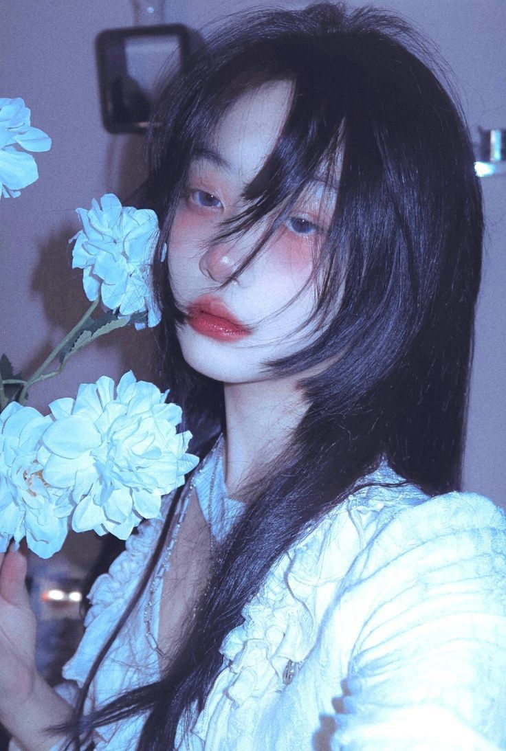 a young woman with long dark hair and rosy cheeks poses for a selfie with soft blue flowers in the foreground. she wears a white lace top and her overall look is delicate and feminine. the image has a soft, ethereal quality with a cool color palette.