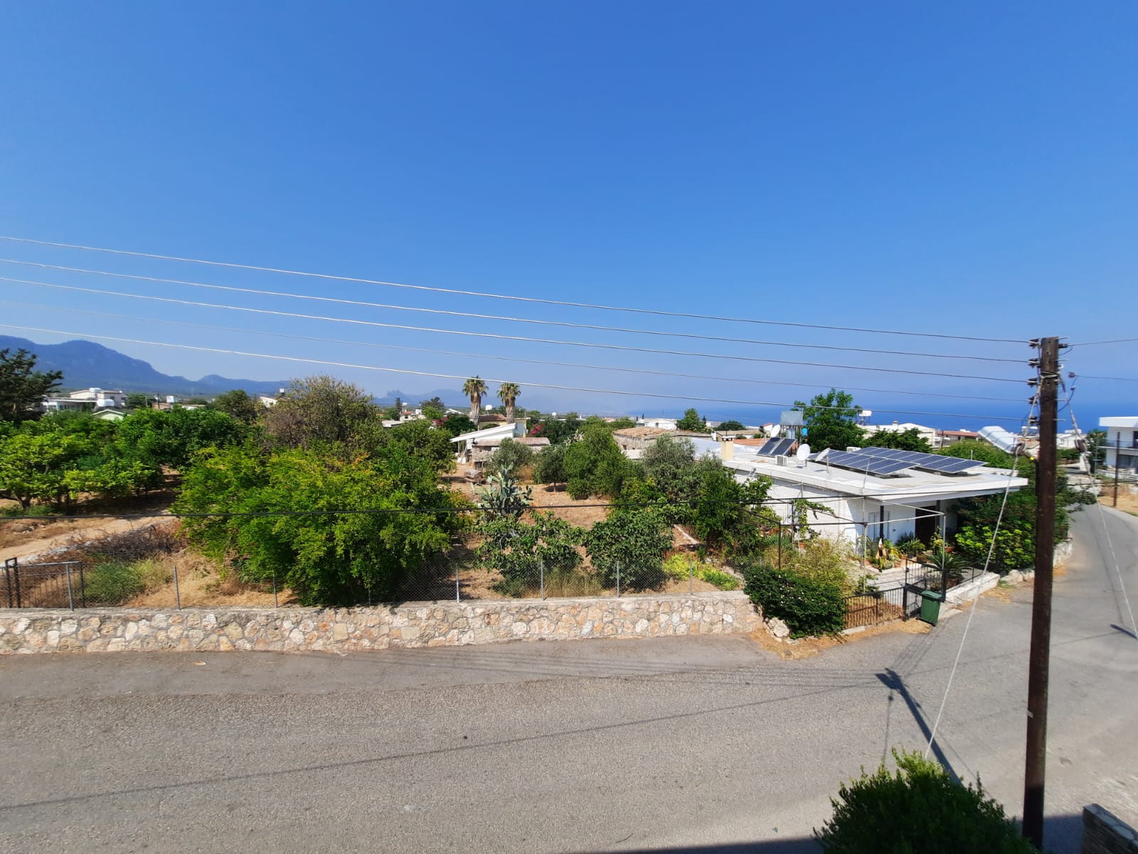a landscape view with multiple houses, green trees, stone wall, road, electric pole, mountains, blue sky, sea, and solar panels on the roof.