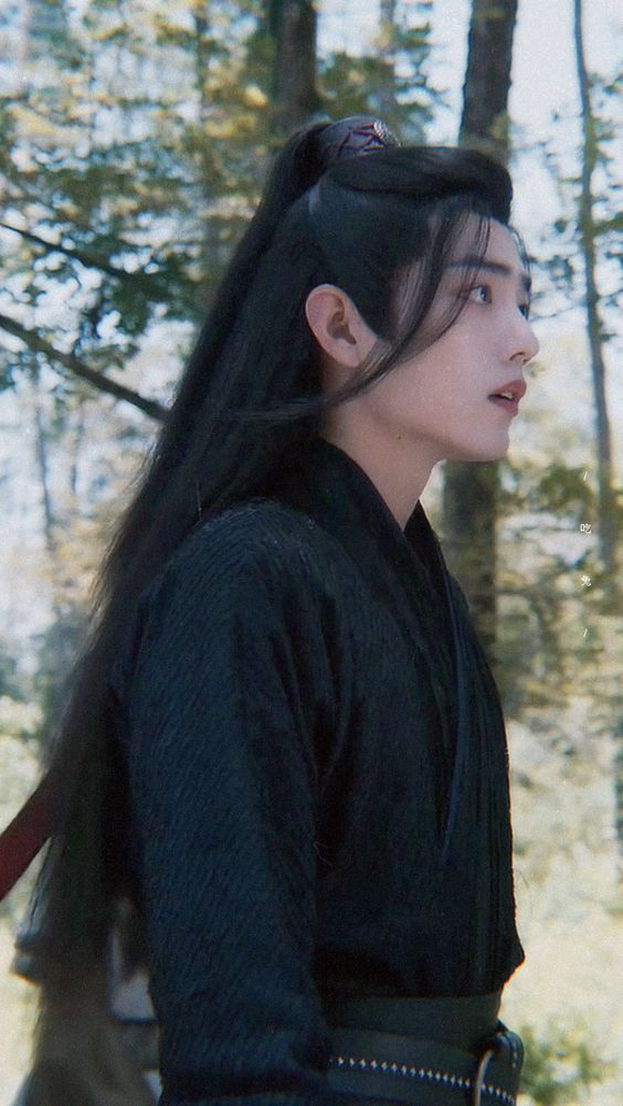 the image shows a young man with long black hair, partially tied back, wearing a dark green hanfu. he is standing in a forest, his head turned to the right, as if looking at something in the distance. the environment is bright, suggesting daylight and natural lighting. the overall atmosphere is peaceful and serene.