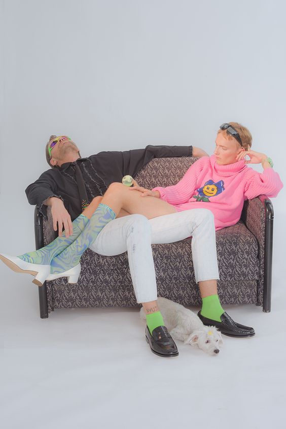 a man and a woman are sitting on a couch. the man is wearing a black shirt and white pants. the woman is wearing a pink sweater and white pants. they both have green socks and black shoes on. there is a white dog sitting in front of them. the man is looking up at the camera and smiling. the woman is looking to the left and smiling. they both seem to be having fun.