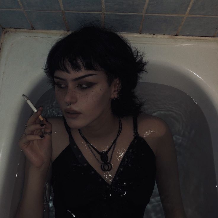 a young woman with black hair and dark makeup is sitting in a bathtub. she is wearing a black tank top, and her hair is wet. she is holding a cigarette in her left hand, and there are rings and a necklace on her fingers and neck. her eyes are looking down. she is surrounded by water in the tub. the water is dark and murky. the overall mood of the image is dark and melancholic.