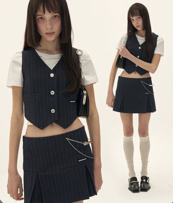 two young women, both with black hair and wearing black pinstripe skirts and vests, pose in a white background studio setting. the woman on the left is wearing a white shirt and a black handbag with a chain strap. the woman on the right is wearing a white shirt and black loafers. both women have on white socks.  their outfits are stylish and trendy.