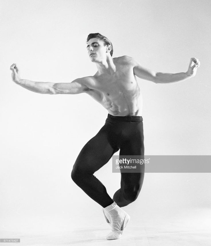 a black and white photo of a shirtless male ballet dancer in a studio. he is wearing black tights and white socks. he is posing with his right arm raised and his left leg bent behind him.