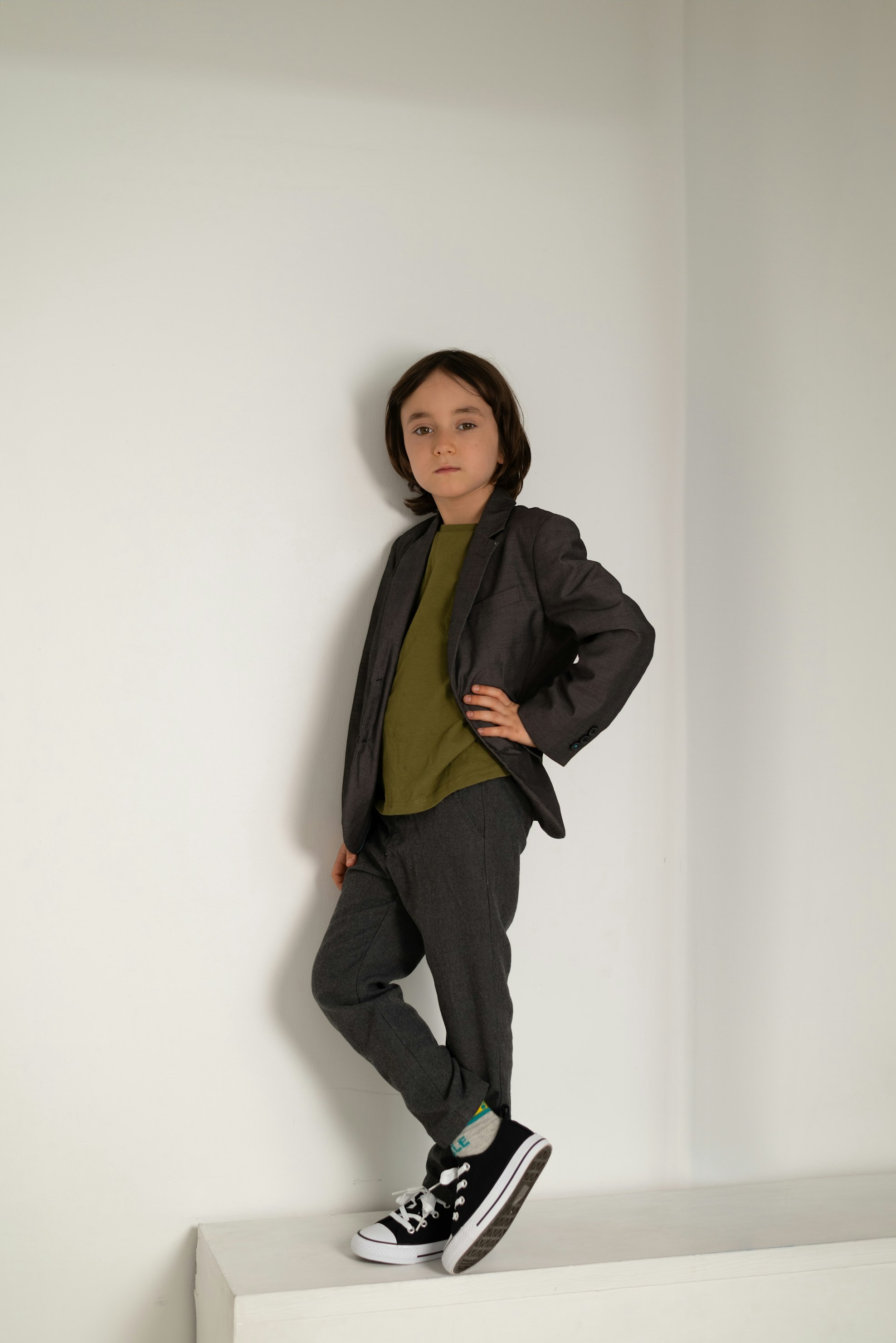 a young boy stands indoors in front of a white wall corner. he is wearing a modern grey suit with a olive green tshirt underneath. the boy is leaning against the wall with one foot placed on a ledge and the other on the floor. he is wearing black and white converse sneakers. the boy is in a fashion portrait pose with one hand on his hip.