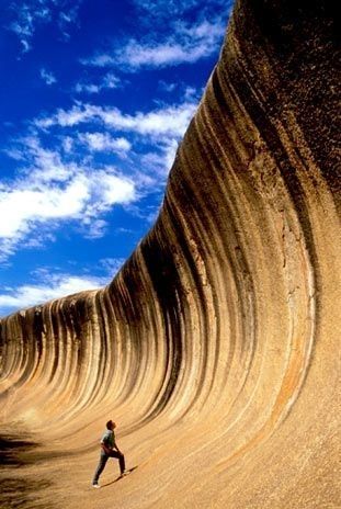 a photo of wave rock, a natural rock formation in western australia. the rock is shaped like a breaking wave, and its surface is covered in intricate striations.  a lone figure stands at the base of the rock, dwarfed by its size.  the sky is a deep blue with fluffy white clouds.  the image is a beautiful example of the power of nature and the wonder of the natural world.
