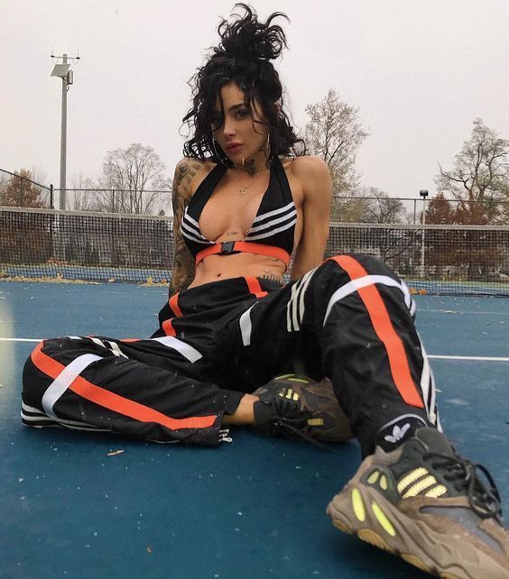 a young woman with tattoos and curly dark hair is posing on a blue tennis court. she is wearing a black and orange athletic outfit, including a sports bra, track pants, and adidas yeezy 700 sneakers. the photo is taken from an eyelevel perspective, and the lighting is overcast and natural.