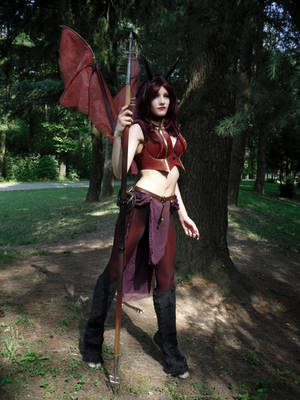 a woman in a fantasy cosplay costume stands in a forest. she is dressed as a demon or succubus with red wings and armor. she holds a spear. the setting is outdoors in a wooded area. the image is welllit and shows detail in the costume. the background is blurred to focus on the subject.