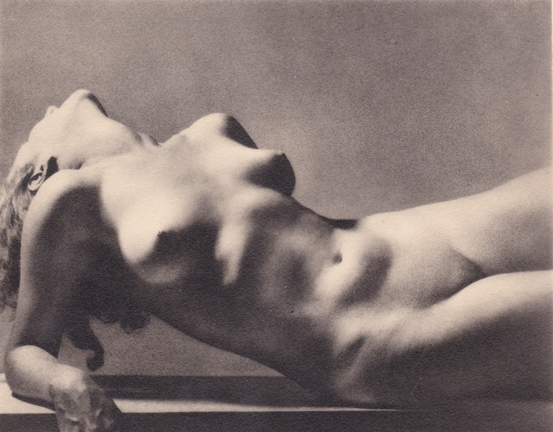 the image is a black and white photograph of a nude female reclining on a surface.  the woman's body is positioned in a graceful arc, her head tilted back. the lighting emphasizes the curves of her body, creating shadows that highlight her form. the texture of the skin is visible, adding to the photo's realism. the overall aesthetic suggests a vintage feel, possibly from the mid20th century. the photograph is artistic in its composition and use of light and shadow to create a sensual and evocative mood.