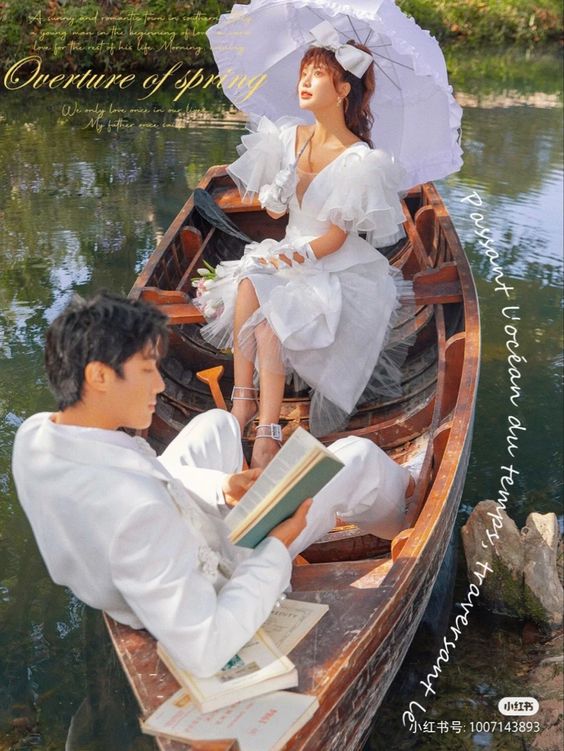 a man and a woman dressed in white are posing in a wooden rowboat on a river. the woman is holding a white parasol and looking up dreamily, while the man is seated and reading a book. the environment suggests a sunny spring day and features lush greenery in the background. the image evokes a sense of romance and celebration, possibly a wedding or engagement photoshoot.