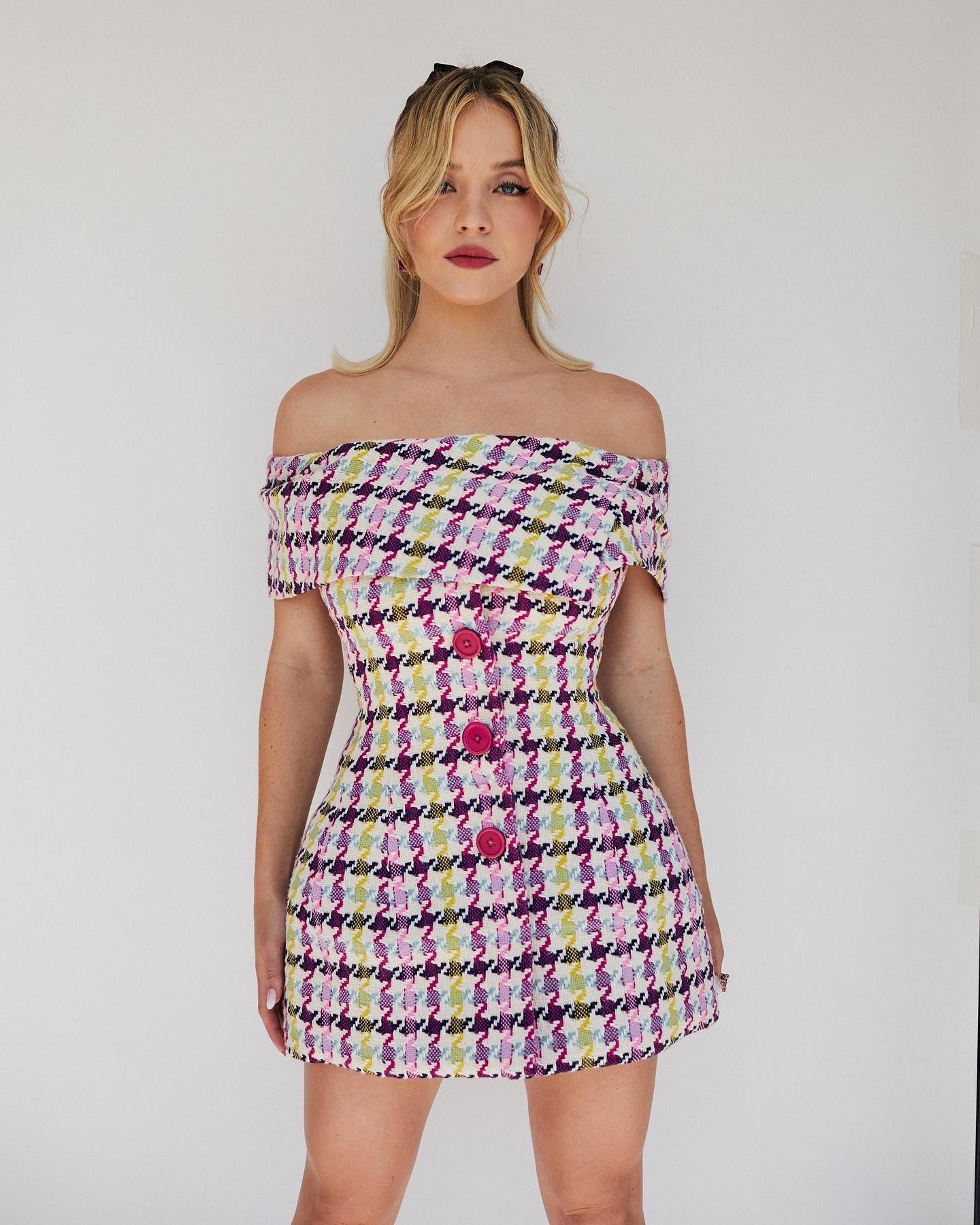 a young blonde woman is standing against a white background, wearing a short, offtheshoulder dress with a houndstooth pattern in shades of pink, purple, green and white. she has her hair styled in soft waves, and she's looking directly at the camera with a confident expression.  the dress is cinched at the waist and flares slightly at the hem.  the buttons on the dress are a bright pink.