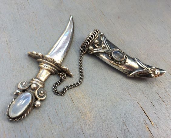 this is a close up shot of two vintage silver sword brooches. they are laid on a wooden surface and appear to have moonstones set in them.