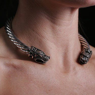 a closeup shot features a silver torc necklace with dragon head terminals worn around a woman's neck. the necklace is made of twisted metal.